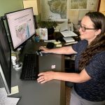 A woman with light skin and long brown hair and glasses working at a desk with dual monitors displaying a detailed map and related data. The workspace includes maps on the wall, papers, and office supplies, indicating a focus on cartography and geology.