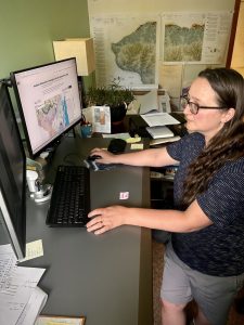 A woman with light skin and long brown hair and glasses working at a desk with dual monitors displaying a detailed map and related data. The workspace includes maps on the wall, papers, and office supplies, indicating a focus on cartography and geology.