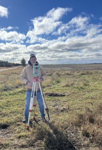 Man in cold weather gear using survey equipment to sight a new well location on a sunny day.