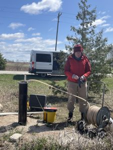 Woman outside in cold weather gear on a sunny day sampling at a well.